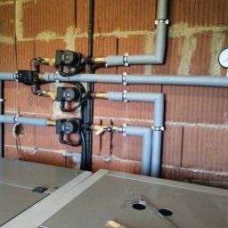 Defro Energy - Szare rury instalacji grzewczej z manometrem i pompami obiegowymi na tle ceglanej ściany w surowym stanie.