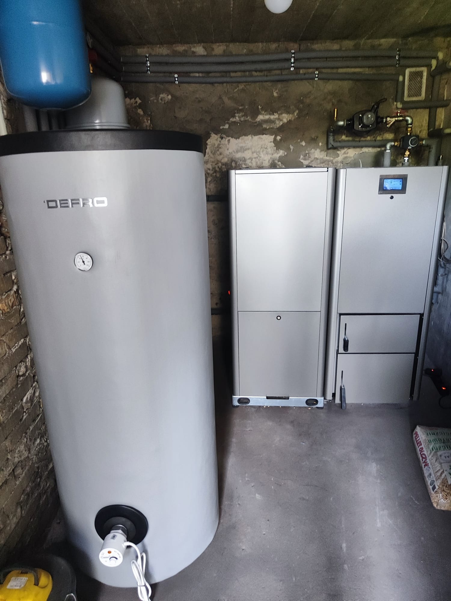 Szary piec z podajnikiem na pellet marki DEFRO w kotłowni, obok widoczny zasobnik na wodę i worki z pelletem.