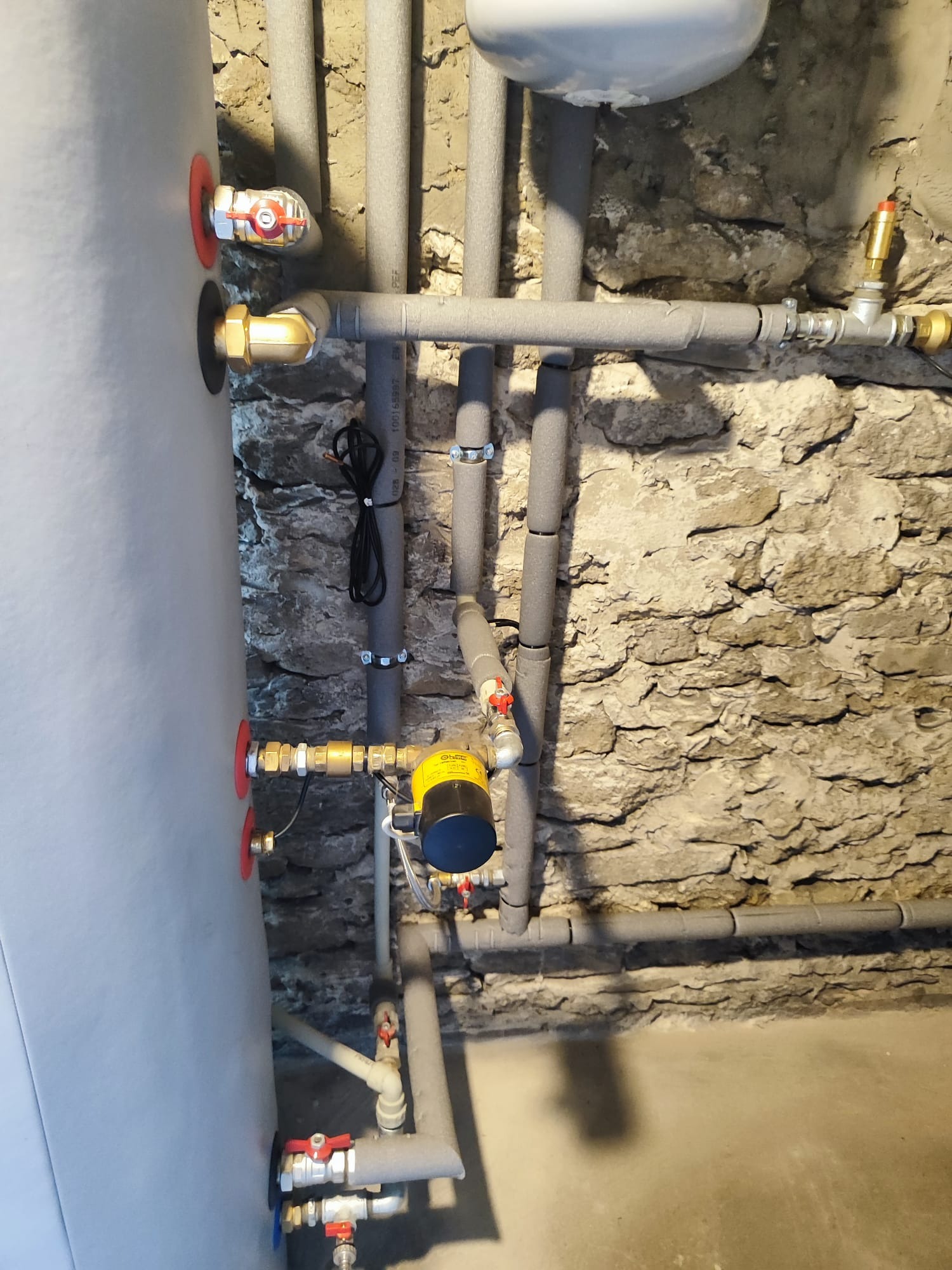 Szary zbiornik akumulacyjny z zaworami i rurami zaizolowanymi termicznie, zamontowany przy kamiennej ścianie w piwnicy w Krakowie.