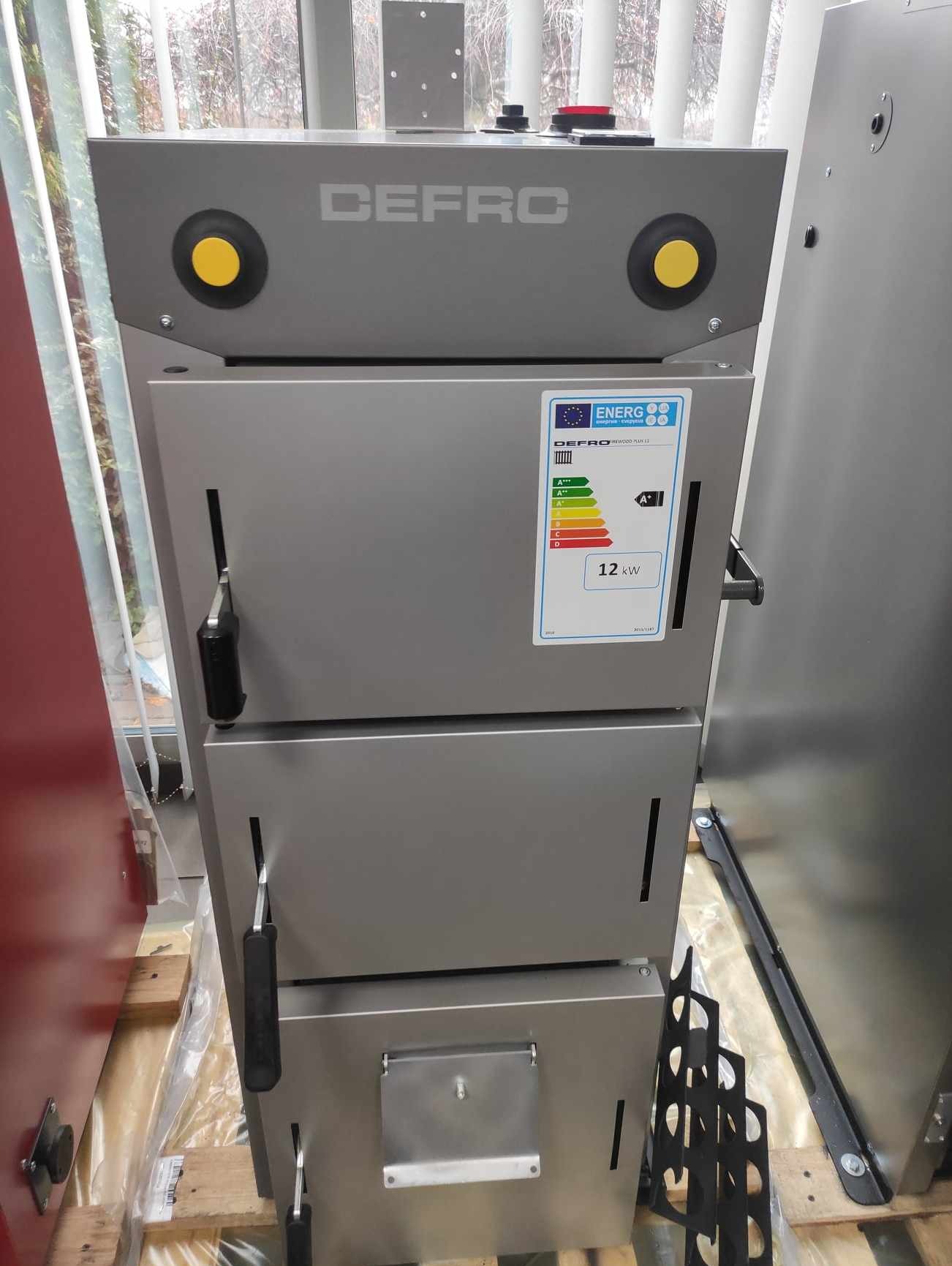 Szary piec marki DEFRO z etykietą energetyczną A+ o mocy 12kW, widoczne czarne uchwyty i żółte przyciski, ustawiony na palecie w pomieszczeniu.