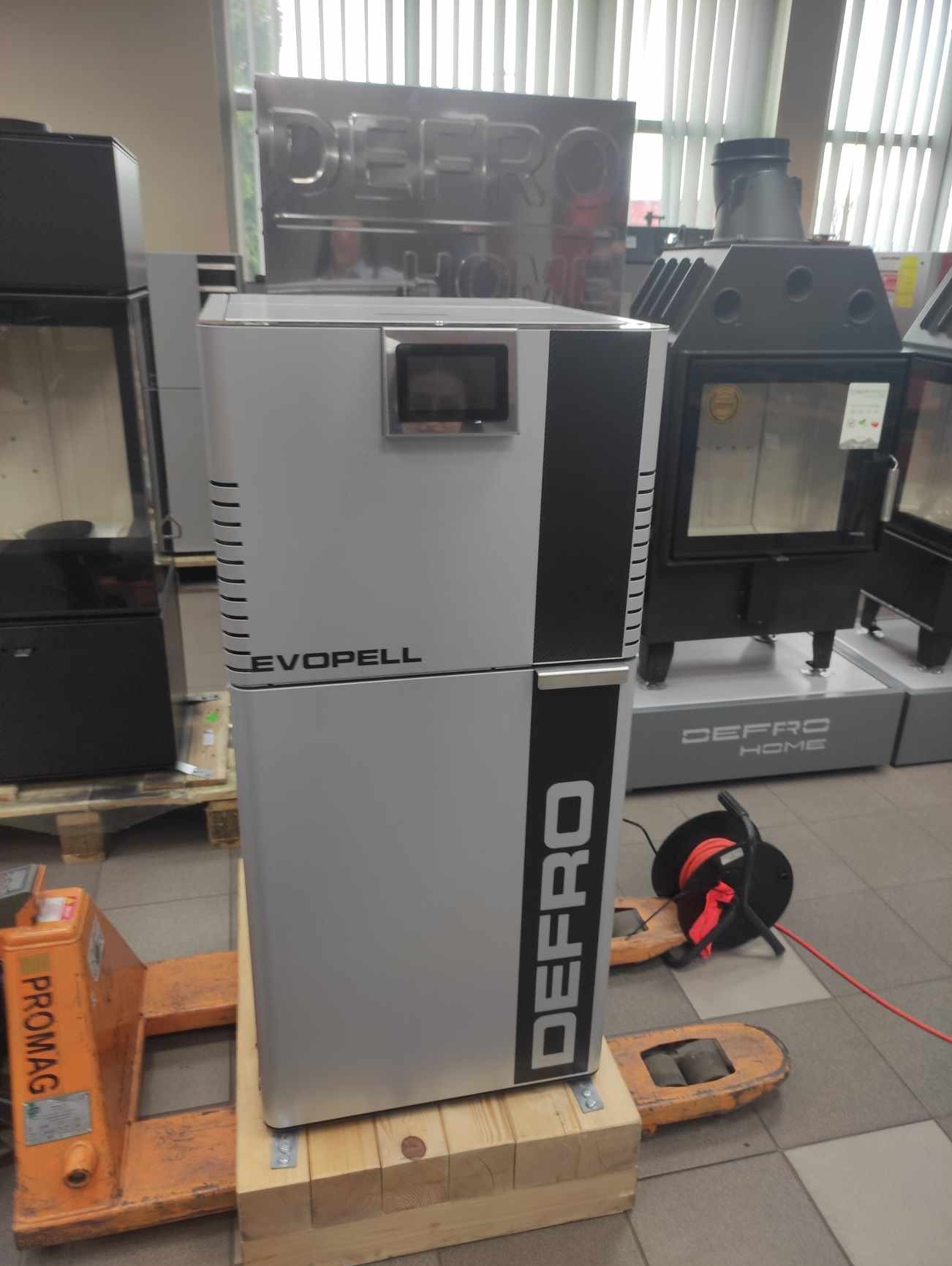 Szary piec na pellet marki Defro Evopell, umieszczony na palecie, w tle inne piece tej samej marki i okno z żaluzjami.