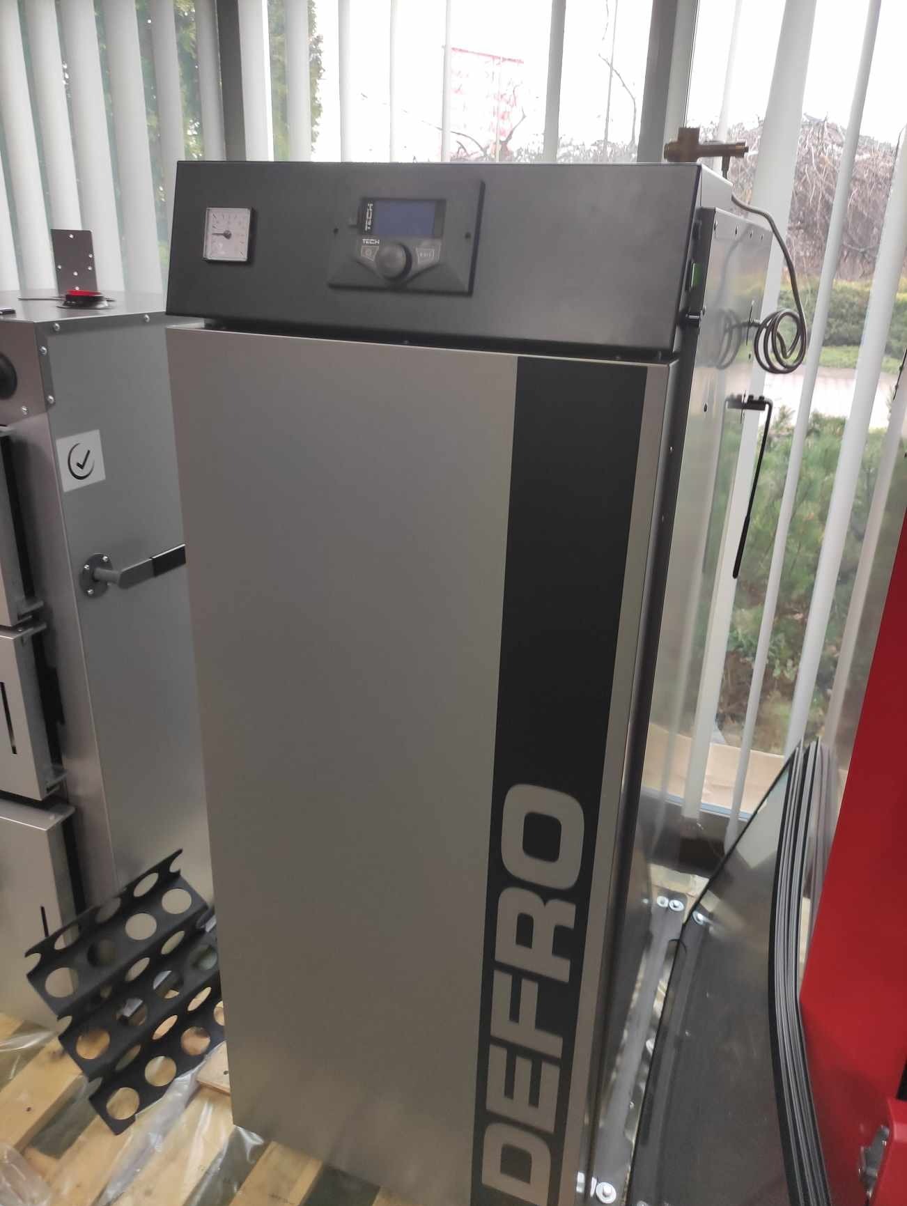Szary piec na paliwo stałe marki DEFRO z panelem sterowania TECH i zegarem analogowym, widoczne elementy montażowe i kratka paleniskowa na palecie, jasne tło okna z żaluzjami.
