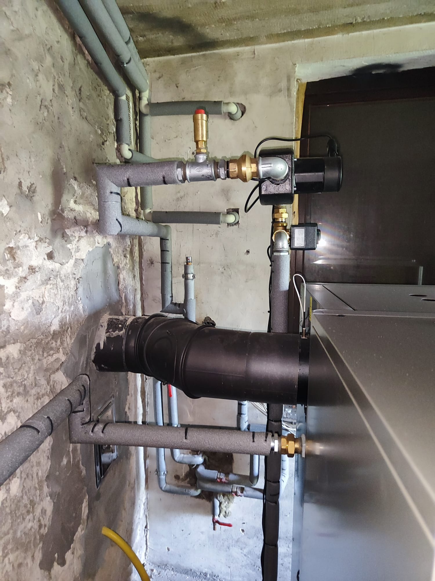 Fragment instalacji grzewczej z rurami w izolacji, zaworami i czarnym przewodem, zamontowanej przy ścianie z widocznymi śladami napraw.