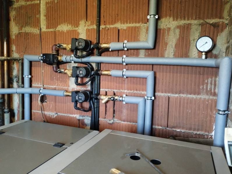 Szare rury instalacji grzewczej z manometrem i pompami obiegowymi na tle ceglanej ściany w surowym stanie.