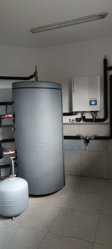 Zamknięte naczynie wzbiorcze i duży, izolowany zasobnik ciepłej wody użytkowej oraz wisząca jednostka sterująca z wyświetlaczem, zainstalowane w pomieszczeniu z wyłożonymi płytkami ścianami i podłogą.