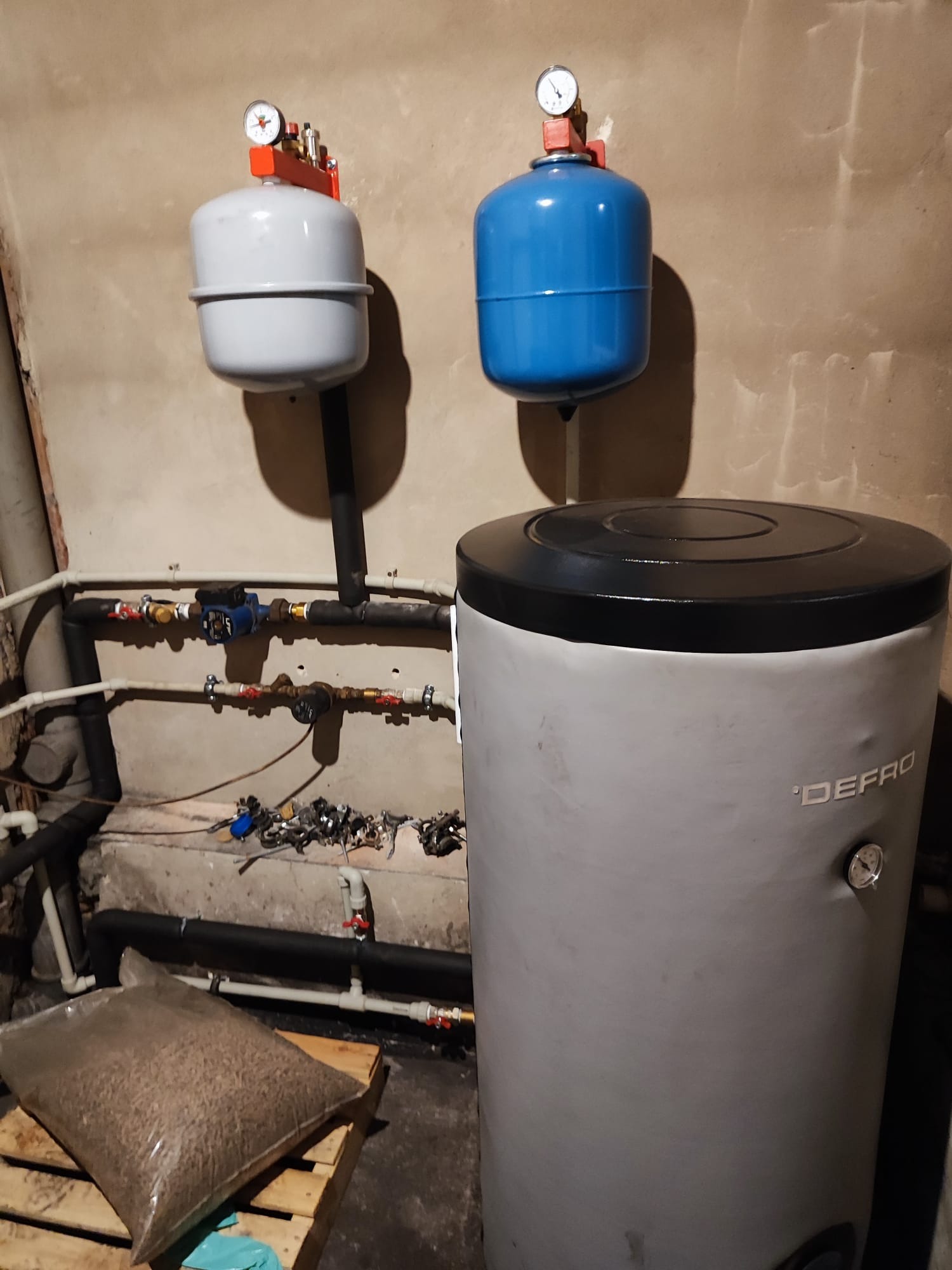 Szary zbiornik buforowy DEFRO z czarną pokrywą, obok worka z pelletem na palecie, z widocznymi rurami i manometrami na ścianie w surowym pomieszczeniu.
