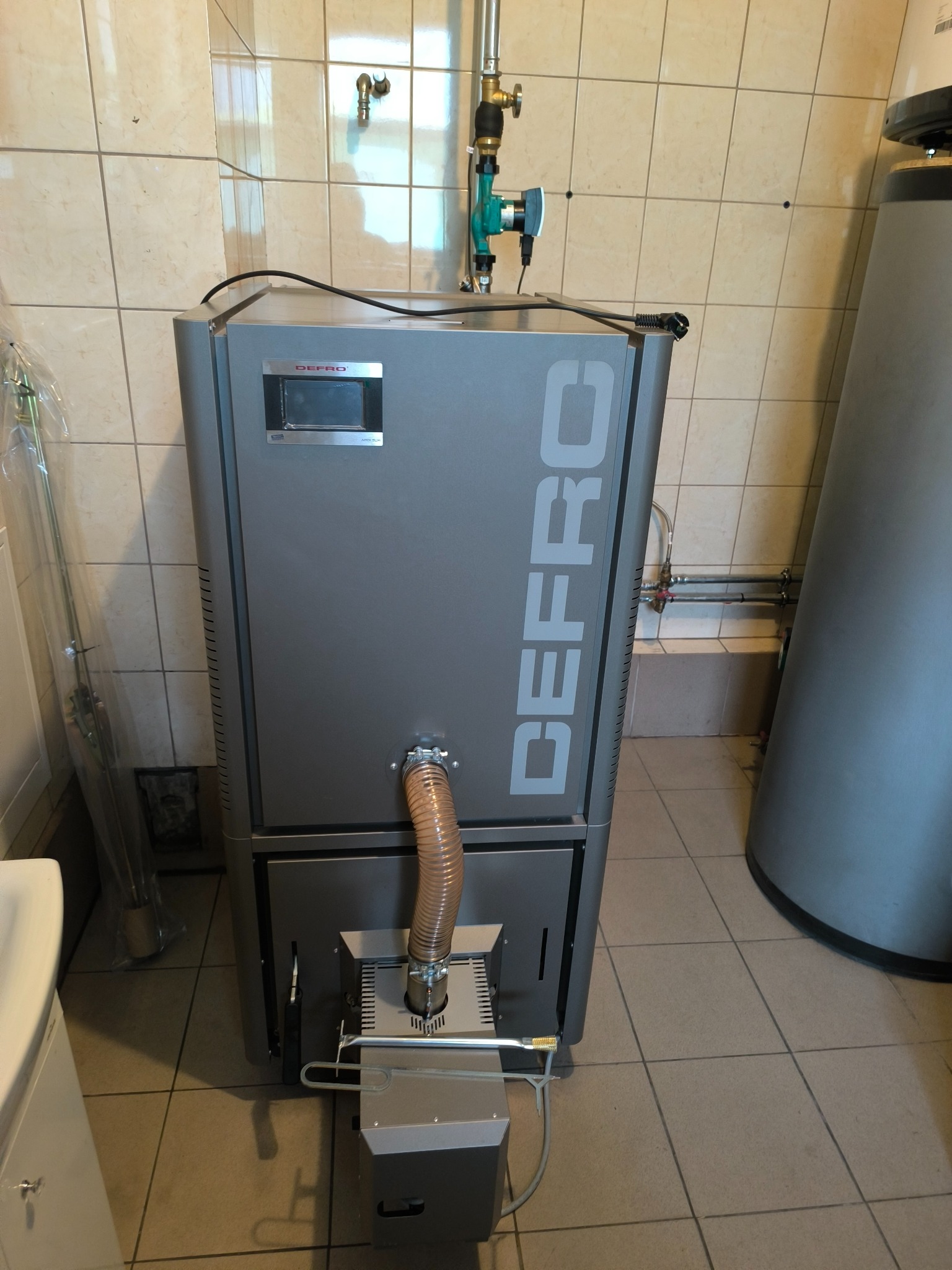 Szary piec marki DEFRO z podłączonymi przewodami i rurą spalinową w kotłowni z beżowymi płytkami, widoczny zasobnik ciepłej wody użytkowej.
