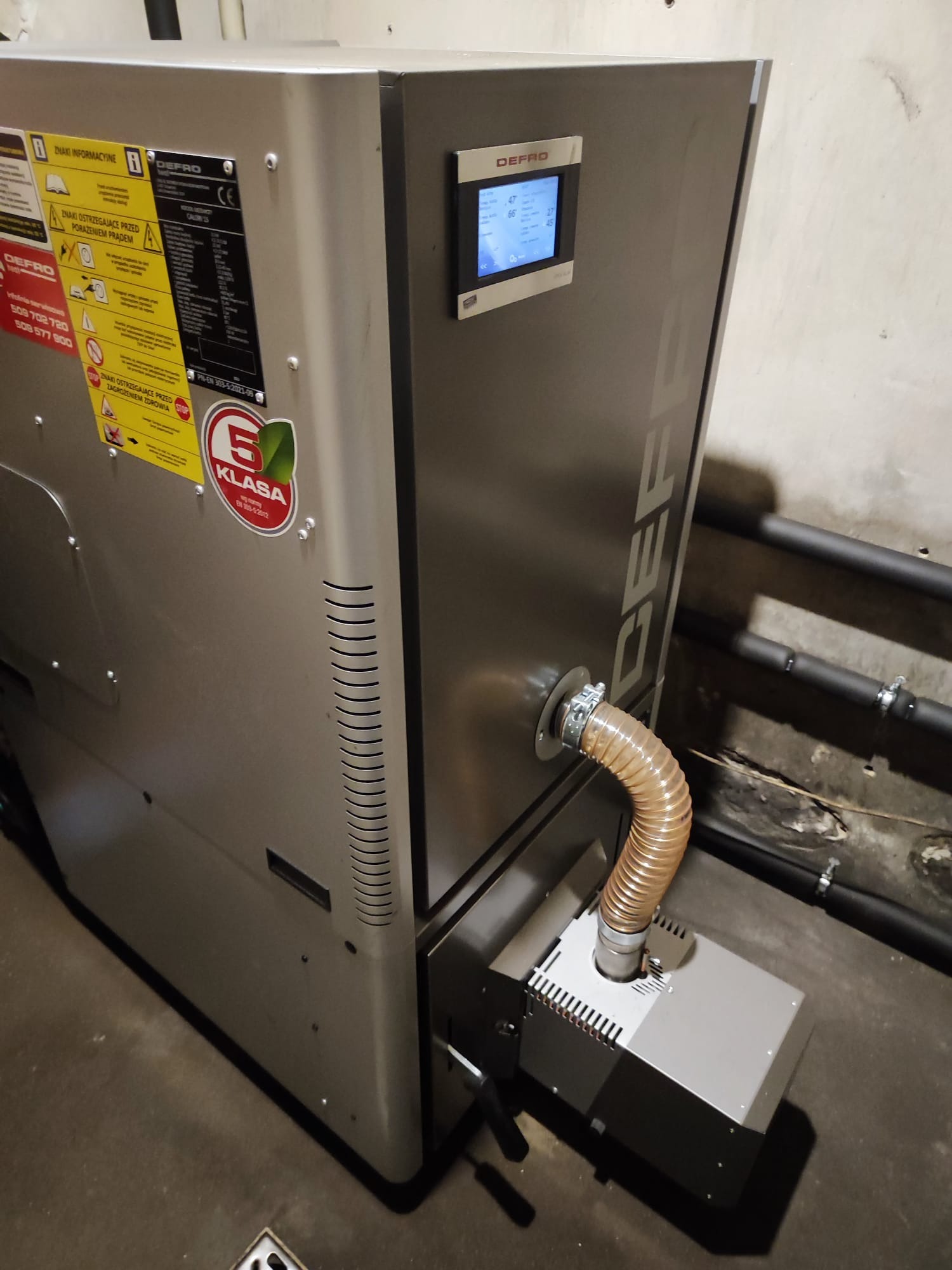 Szary piec na paliwo stałe marki DEFRO z widocznym panelem sterowania, etykietami informacyjnymi i rurą doprowadzającą powietrze do paleniska, umieszczony w pomieszczeniu z rurami grzewczymi w tle.