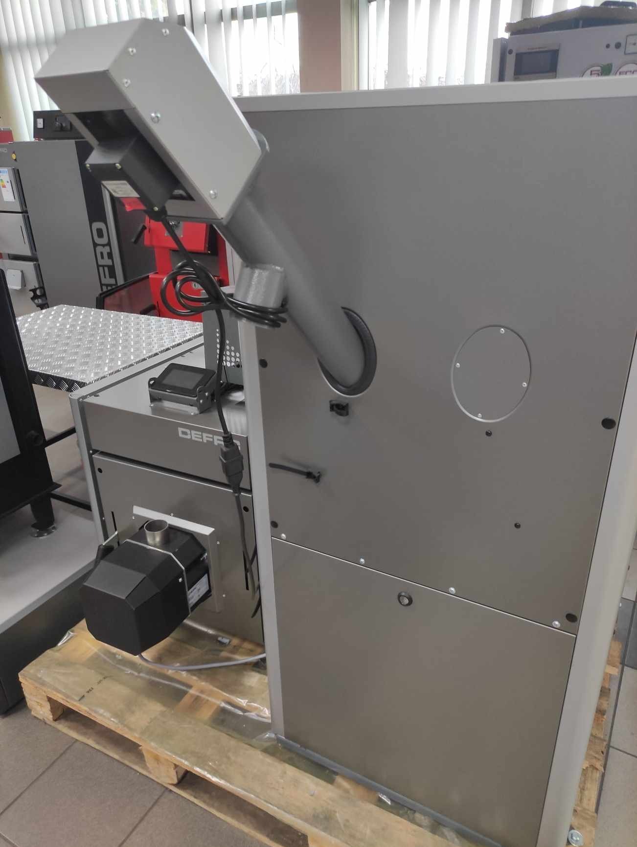 Szary piec z podajnikiem na palecie, widoczny panel sterowania i podajnik paliwa, z boku widoczna etykieta producenta Defro.