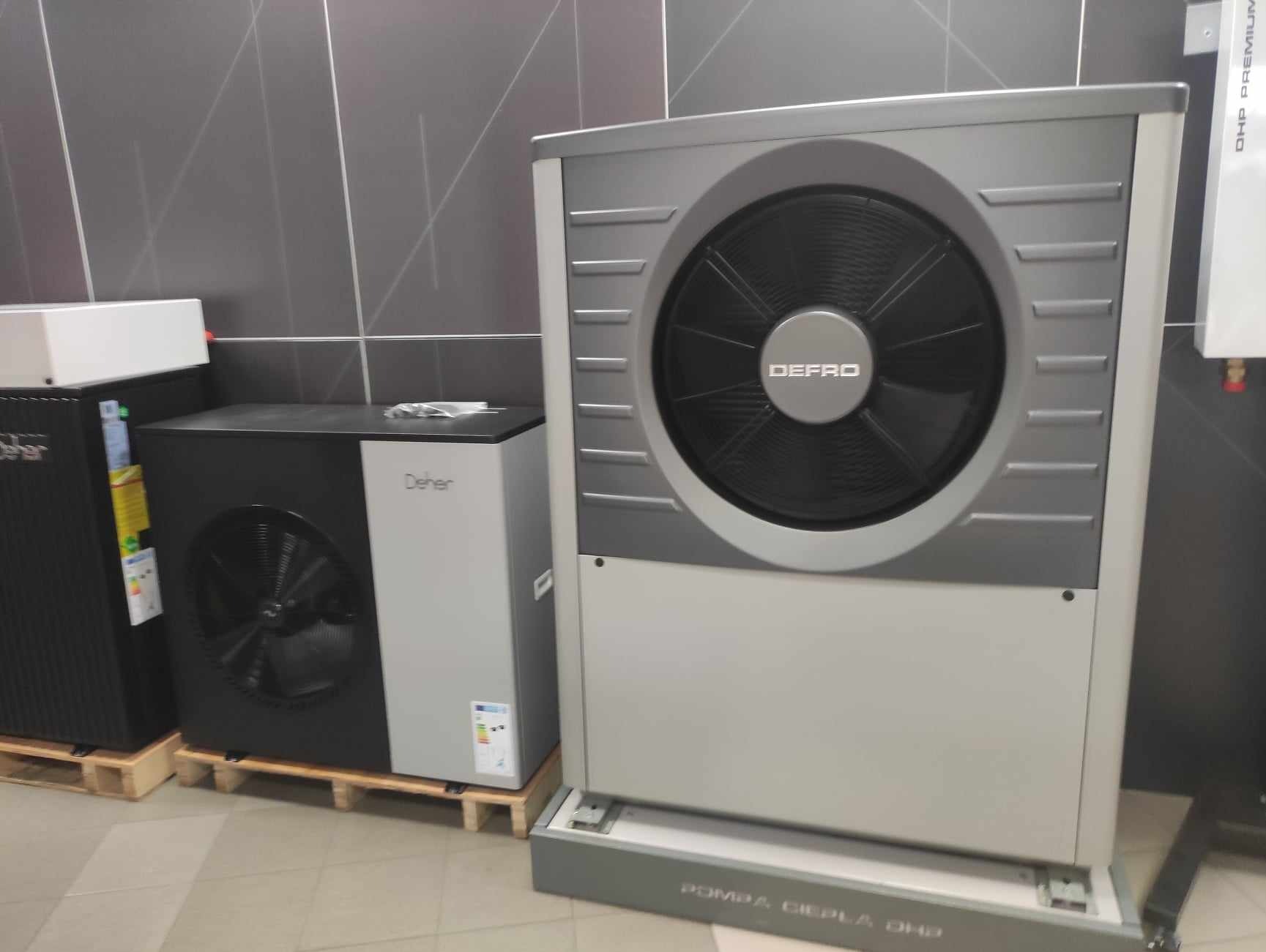 Dwie szare jednostki grzewcze marki Defro, jedna większa z widocznym wentylatorem, druga mniejsza, stojące na drewnianych paletach w pomieszczeniu z szarą płytkową ścianą.