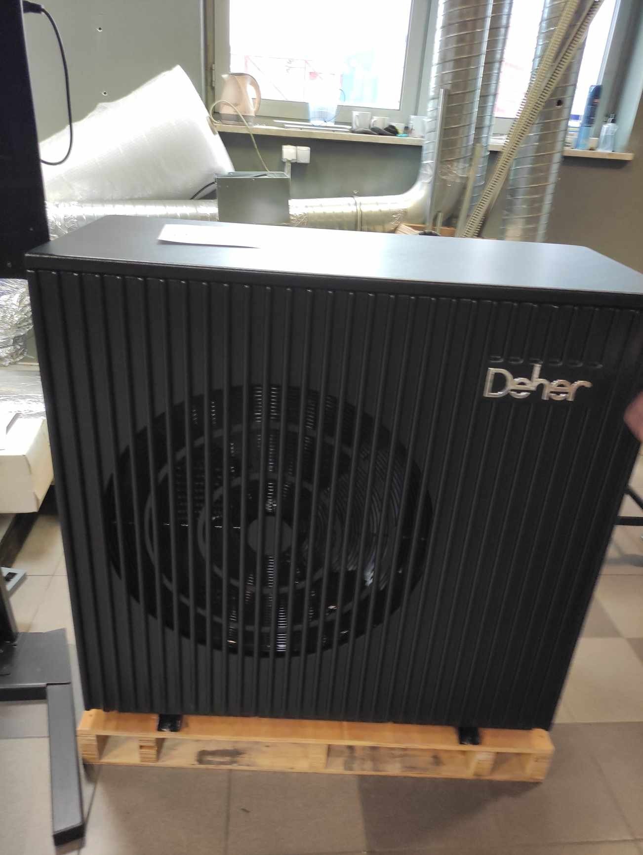 Czarna pompa ciepła marki Deher, stojąca na drewnianej palecie w pomieszczeniu z widocznymi rurami wentylacyjnymi i oknem w tle.