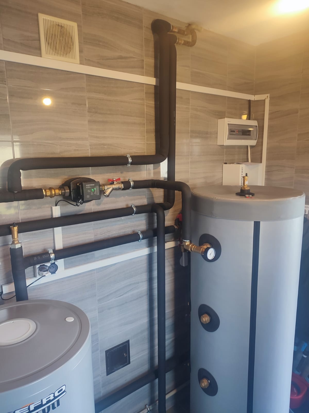 Szare, pionowe zbiorniki na wodę z czarnymi, izolowanymi rurami i armaturą mosiężną na tle płytek imitujących marmur. Widoczny również zawór trójdrożny z elektronicznym sterowaniem.