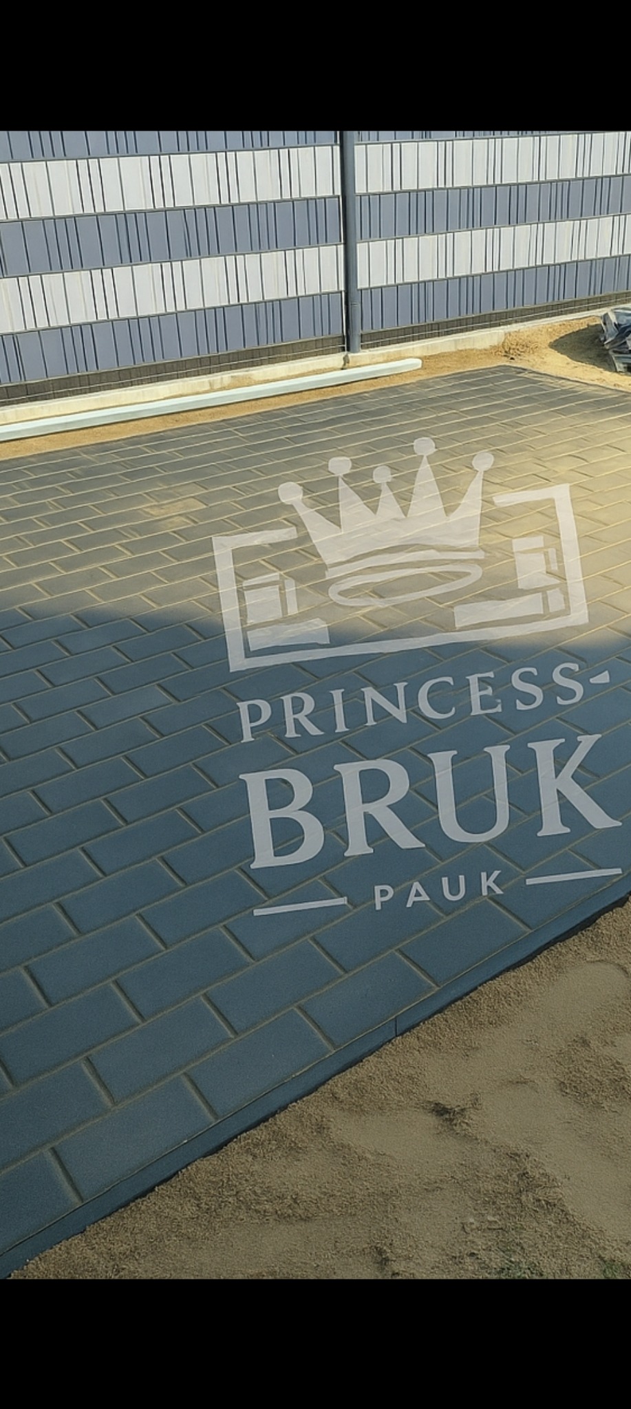 Niebieska kostka brukowa ułożona wzorem jodełki, obok widoczny fragment ogrodzenia panelowego w odcieniach szarości i piasek. Na kostce logo firmy.