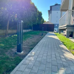 Oświetlona ścieżka z kostki brukowej przy bloku mieszkalnym z nowoczesnymi lampami parkowymi w kształcie prostopadłościanów, widoczne drzewa i budynek w tle.