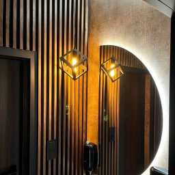 EL-AMPI JAKUB KOWALEWSKI - Nowoczesna łazienka z podświetlanym lustrem w kształcie półksiężyca, drewnianą ścianą z lamelami i designerską lampą wiszącą w formie sześcianu.