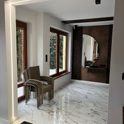 EL-AMPI JAKUB KOWALEWSKI - Nowoczesne wnętrze z marmurową podłogą, oświetleniem LED wzdłuż framugi i dekoracyjnym lustrem na ścianie z drewnianymi lamelami. Stos krzeseł przy oknie.