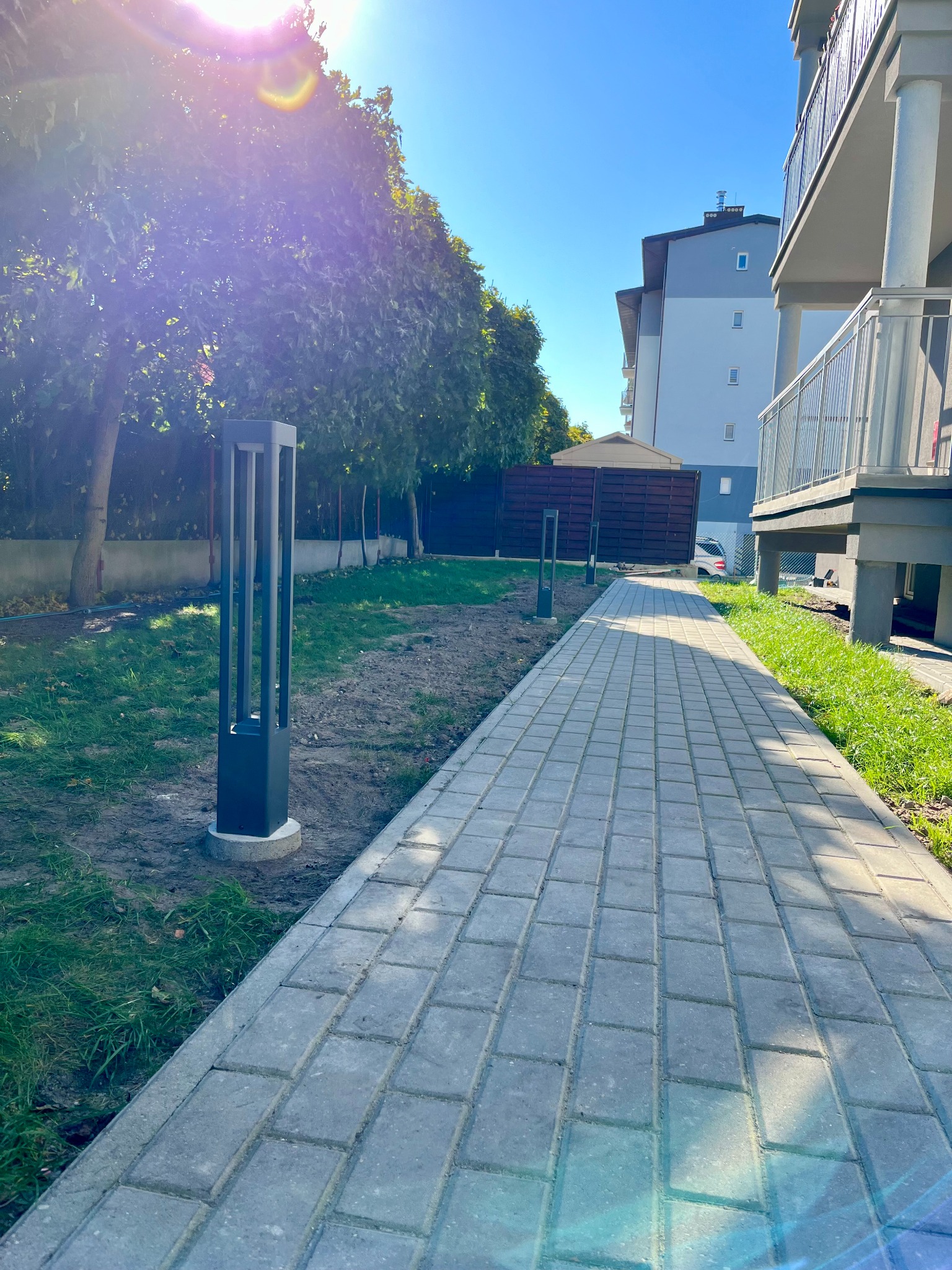 Oświetlona ścieżka z kostki brukowej przy bloku mieszkalnym z nowoczesnymi lampami parkowymi w kształcie prostopadłościanów, widoczne drzewa i budynek w tle.