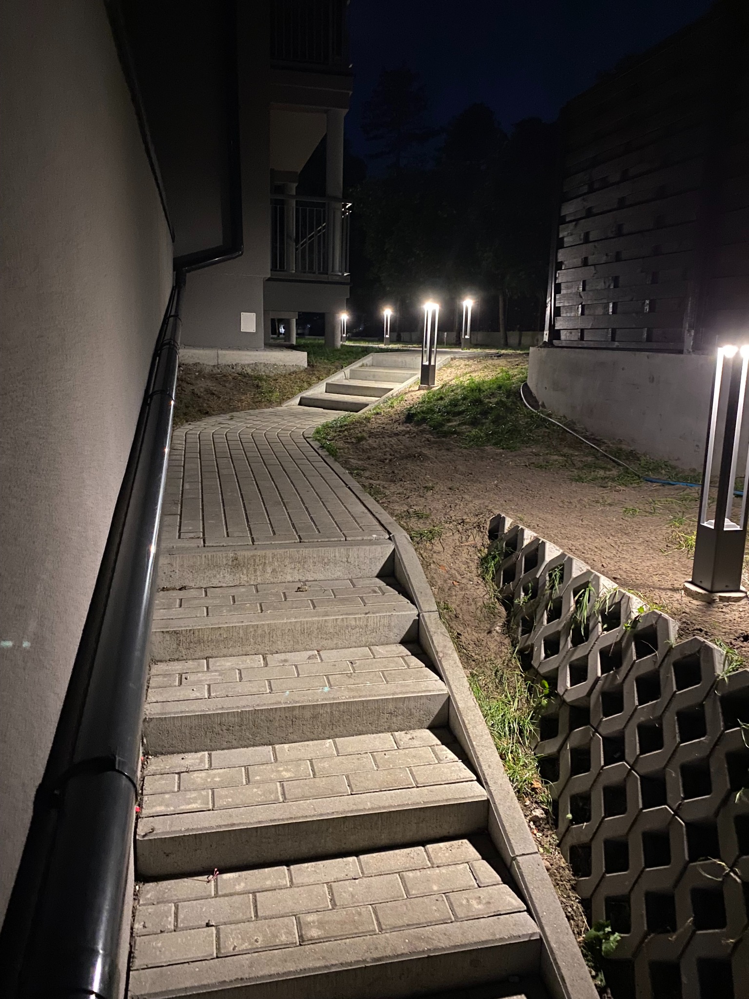 Oświetlona ścieżka z kostki brukowej prowadząca do bloku mieszkalnego nocą. Lampy parkowe oświetlają drogę wzdłuż schodów i muru oporowego z ażurowych betonowych elementów.