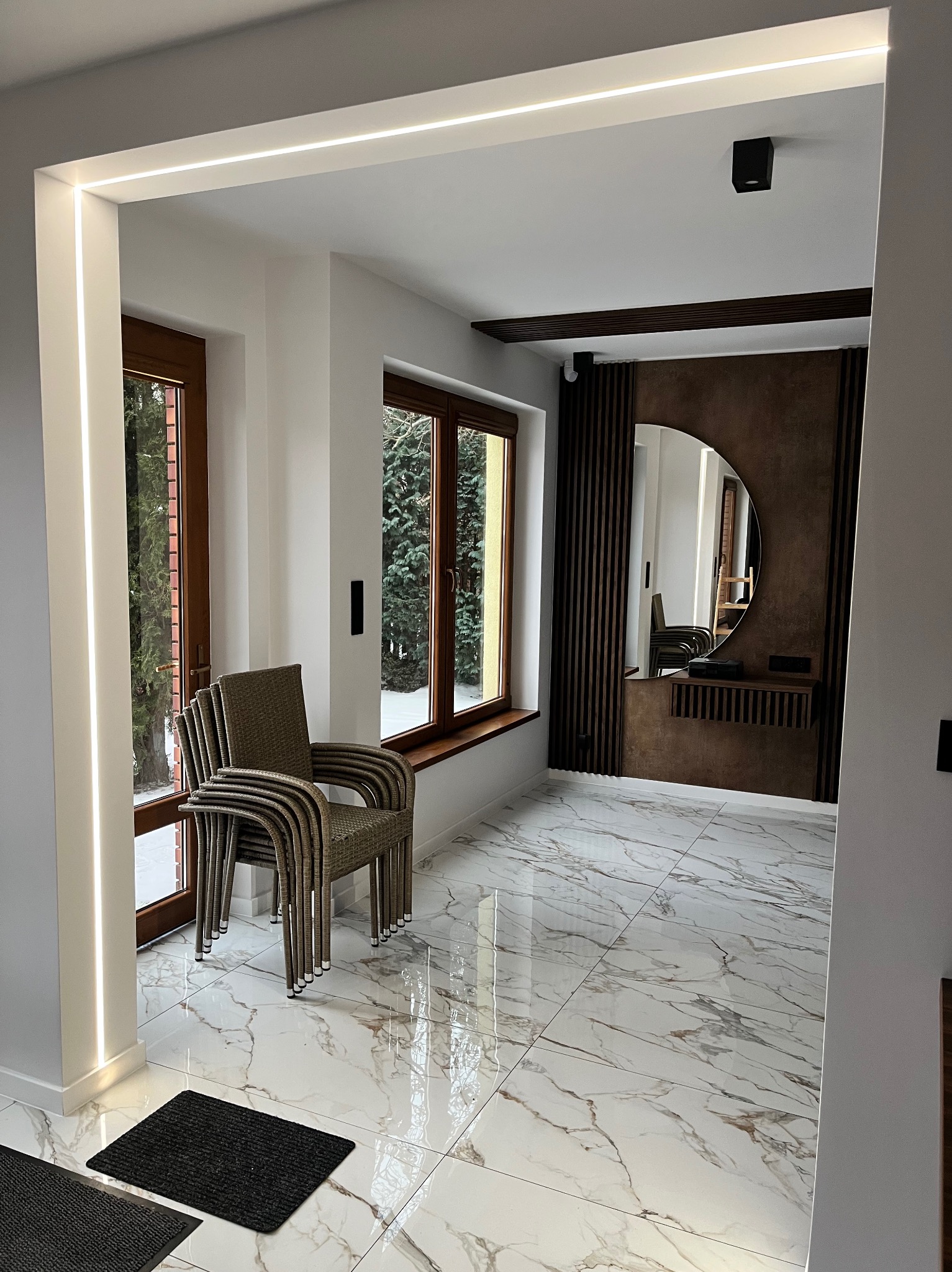 Nowoczesne wnętrze z marmurową podłogą, oświetleniem LED wzdłuż framugi i dekoracyjnym lustrem na ścianie z drewnianymi lamelami. Stos krzeseł przy oknie.