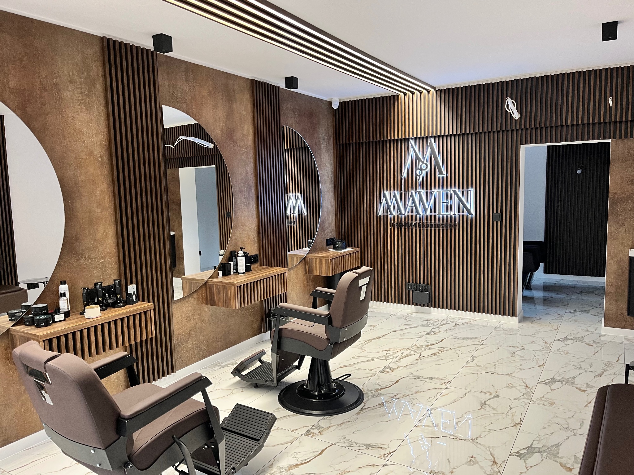 Nowoczesne wnętrze salonu barberskiego z dwoma fotelami, okrągłymi lustrami i drewnianymi panelami ściennymi. Oświetlenie LED podkreśla marmurową podłogę.