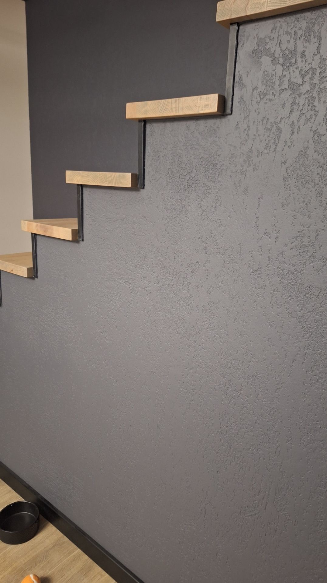 Minimalistyczne schody wspornikowe z drewnianymi stopniami i czarnymi wspornikami na tle szarej ściany o fakturze baranka. W tle widoczny fragment beżowej ściany.