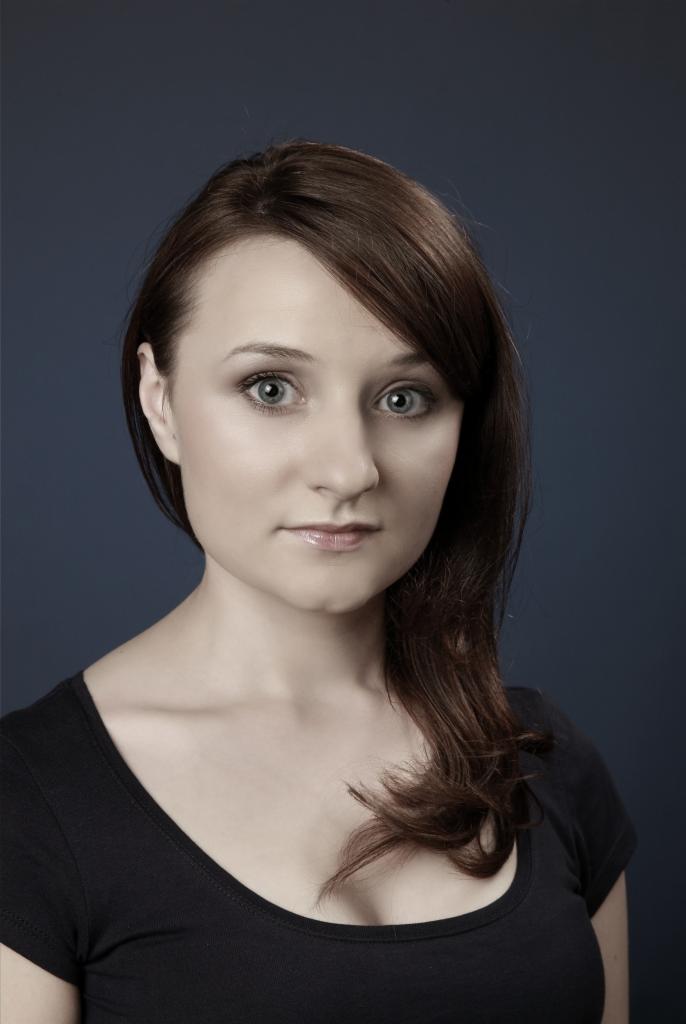 Portret kobiety z brązowymi włosami i niebieskimi oczami na ciemnoniebieskim tle, ubrana w czarną koszulkę.