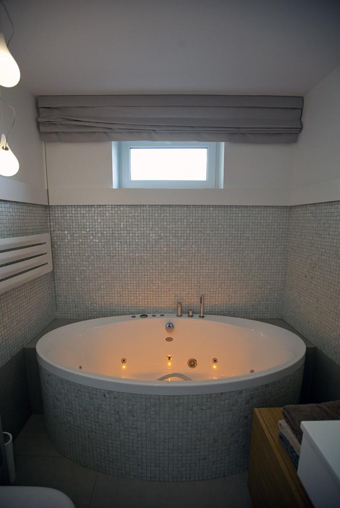 Okrągła wanna z hydromasażem, obudowana mozaiką w odcieniach szarości. Nad wanną okno z szarą roletą rzymską. Ściany wyłożone mozaiką, oświetlenie punktowe.