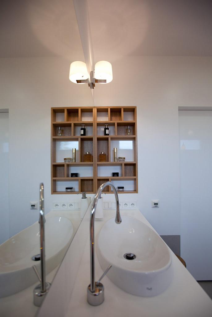 Nowoczesna łazienka z podwójną umywalką, drewnianą półką na kosmetyki i minimalistycznym oświetleniem. Białe ściany i armatura tworzą spójną, elegancką przestrzeń.