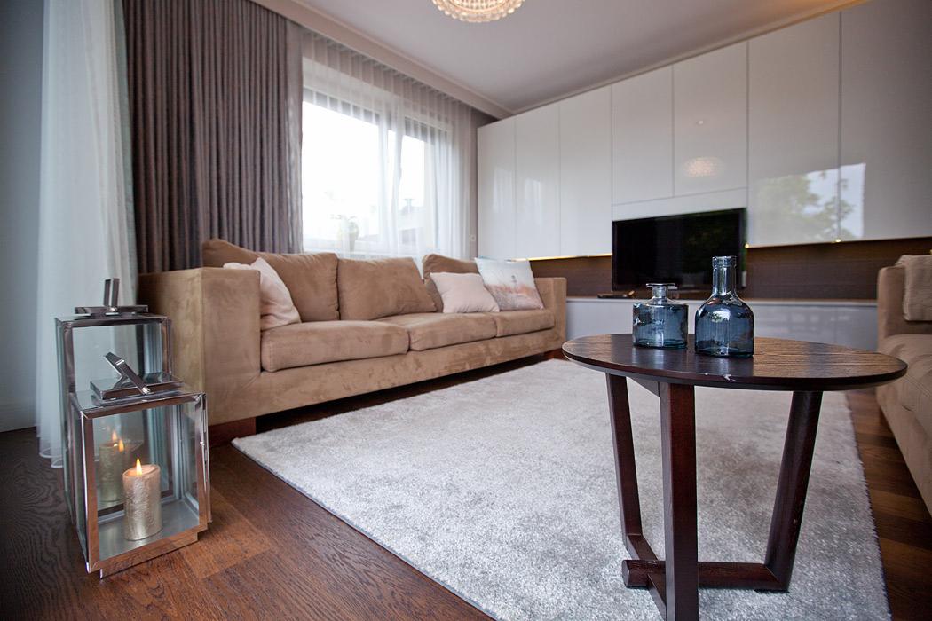 Jasny salon z beżową sofą, drewnianym stolikiem i dekoracyjnymi lampionami. Minimalistyczne białe szafki na ścianie i duże okno z zasłonami tworzą przytulną atmosferę.