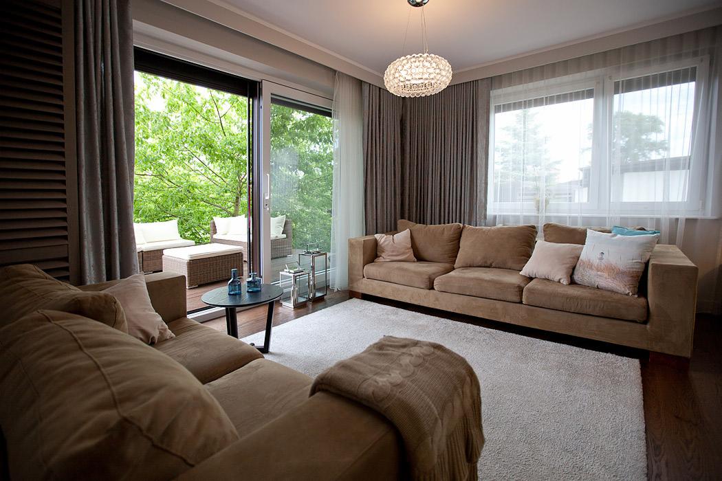 Jasny salon z brązowymi sofami i wyjściem na taras z meblami ogrodowymi. Okno z firankami i lampa wisząca dopełniają aranżację.