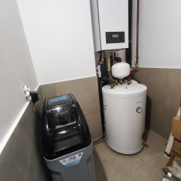 Z Grubej Rury Usługi Hydrauliczne Mariusz Żychowski - Nowoczesna instalacja grzewcza z białym piecem gazowym Brötje, zasobnikiem Trinity i szarym zmiękczaczem Water Technic, w narożniku pomieszczenia z beżowymi płytkami.