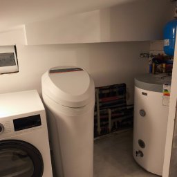 Z Grubej Rury Usługi Hydrauliczne Mariusz Żychowski - Zainstalowany biały zmiękczacz wody Viessmann Aquahome 20 Smart obok pralko-suszarki Bosch Serie 6 i bojlera, z widocznymi rurami hydraulicznymi w tle, w kompaktowej wnęce.