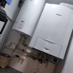 Z Grubej Rury Usługi Hydrauliczne Mariusz Żychowski - Biały piec gazowy Junkers i bojler Ariston z czerwoną diodą, zamontowane na białej ścianie. Widoczne miedziane i plastikowe rury, zawory oraz panel sterujący z zielonymi diodami.