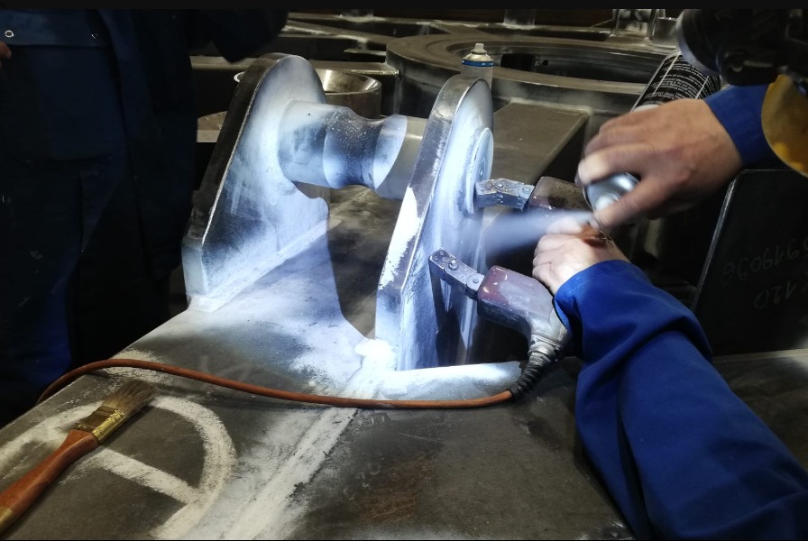 Szlifowanie metalowego elementu konstrukcyjnego za pomocą szlifierki pneumatycznej i preparatu w sprayu, widoczny pędzel i fragmenty odzieży roboczej.