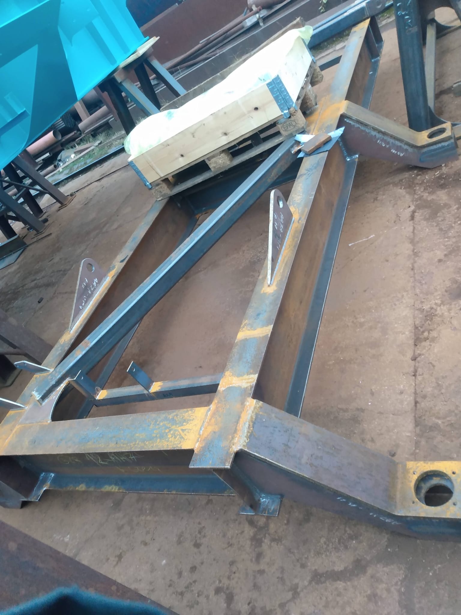 Konstrukcja spawana z metalowych profili o rdzawym nalocie, częściowo pomalowana na niebiesko, leżąca na betonowej posadzce warsztatu, z paletą drewnianą na górze.