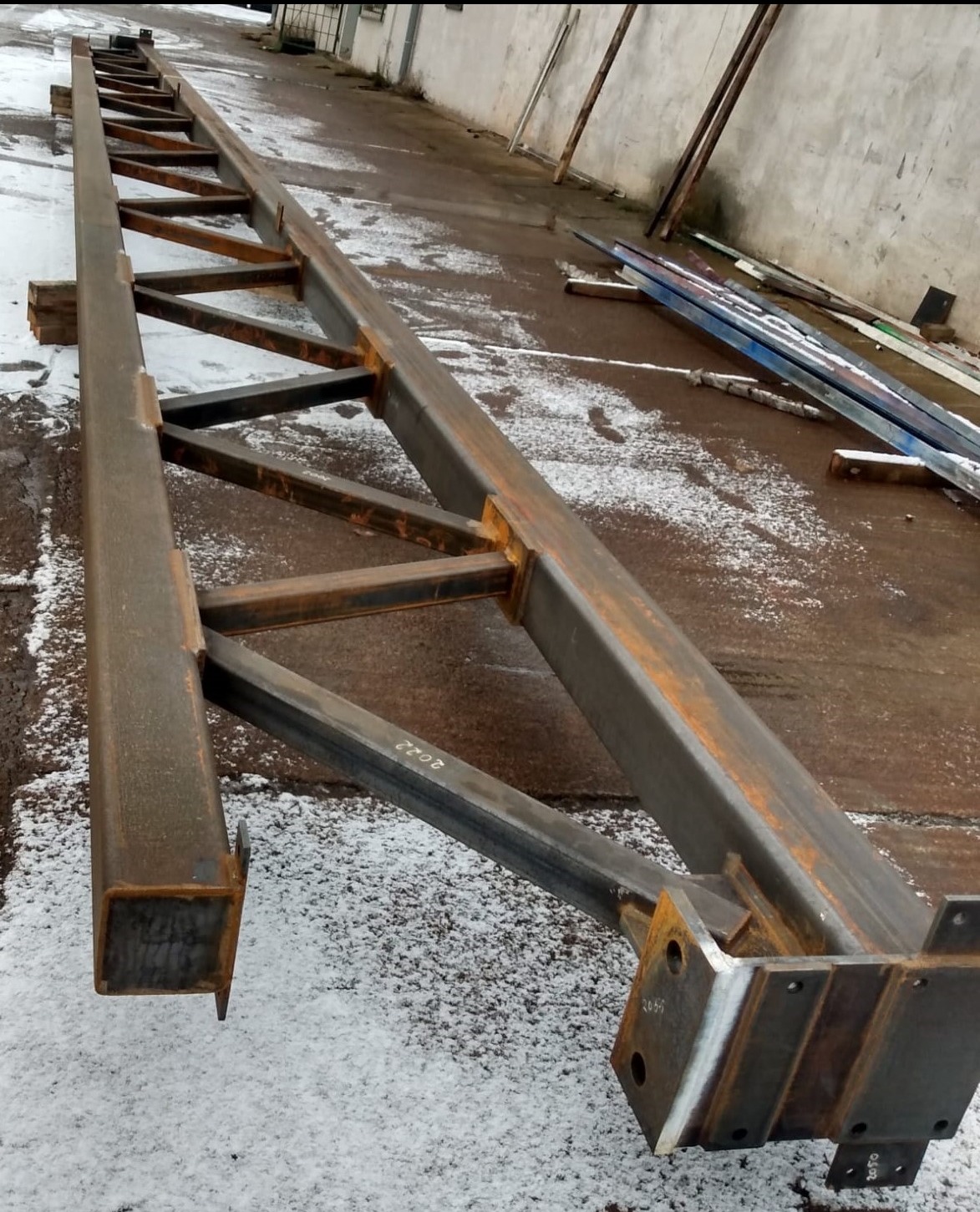 Długa, metalowa konstrukcja kratownicowa leżąca na ziemi z resztkami śniegu, widoczne ślady spawania i rdzy.