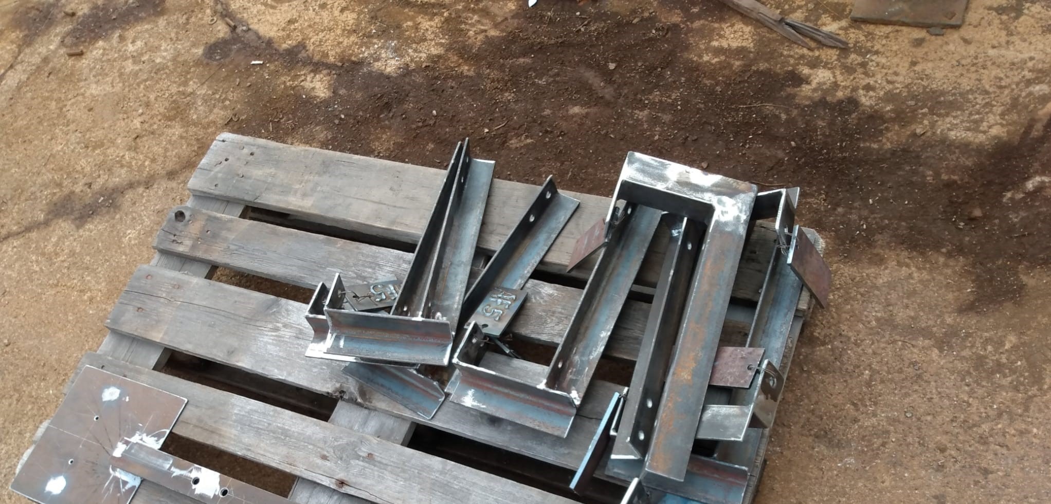 Surowe, stalowe elementy konstrukcyjne, prawdopodobnie do ogrodzenia lub bramy, leżące na drewnianej palecie na tle ziemi.