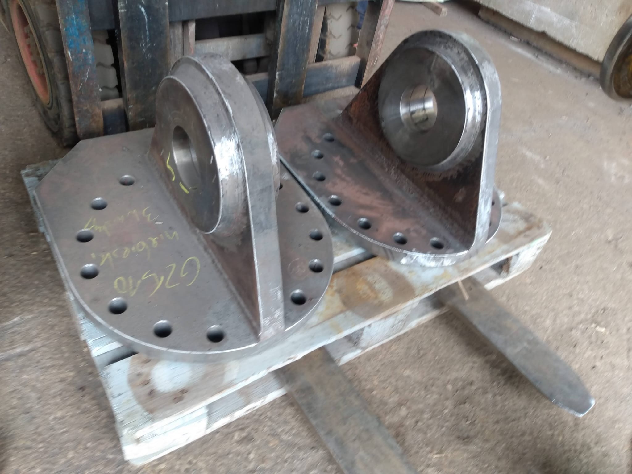 Dwa stalowe elementy konstrukcyjne z okrągłymi otworami, leżące na palecie, widoczne ślady obróbki i spawania.