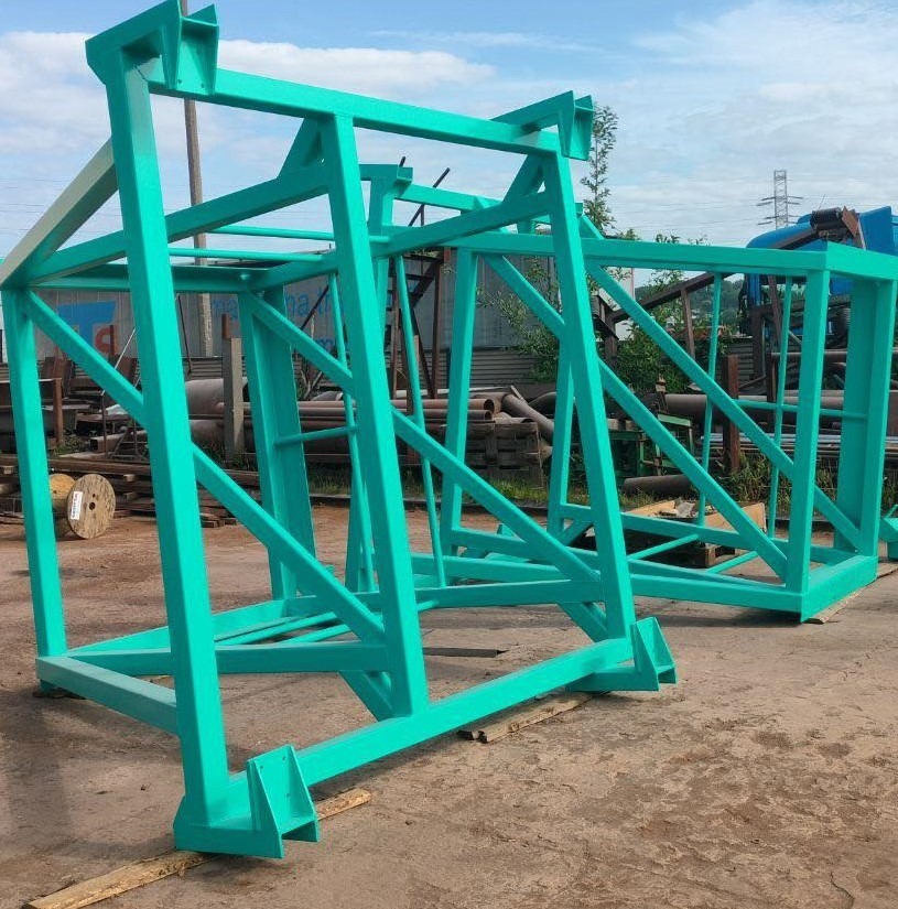 Dwie pomalowane na turkusowo, metalowe konstrukcje szkieletowe, prawdopodobnie bramy lub fragmenty większych konstrukcji, leżące na betonowej powierzchni w otoczeniu rur i innych elementów metalowych.