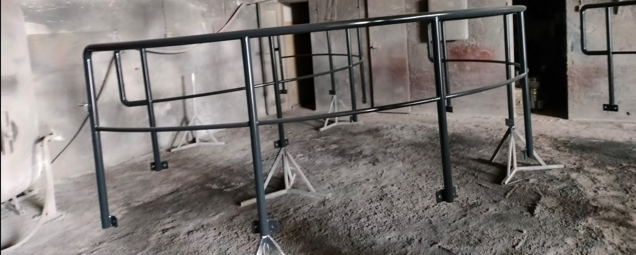 Szare, metalowe barierki na tymczasowych podporach w surowym pomieszczeniu, prawdopodobnie warsztacie lub magazynie.