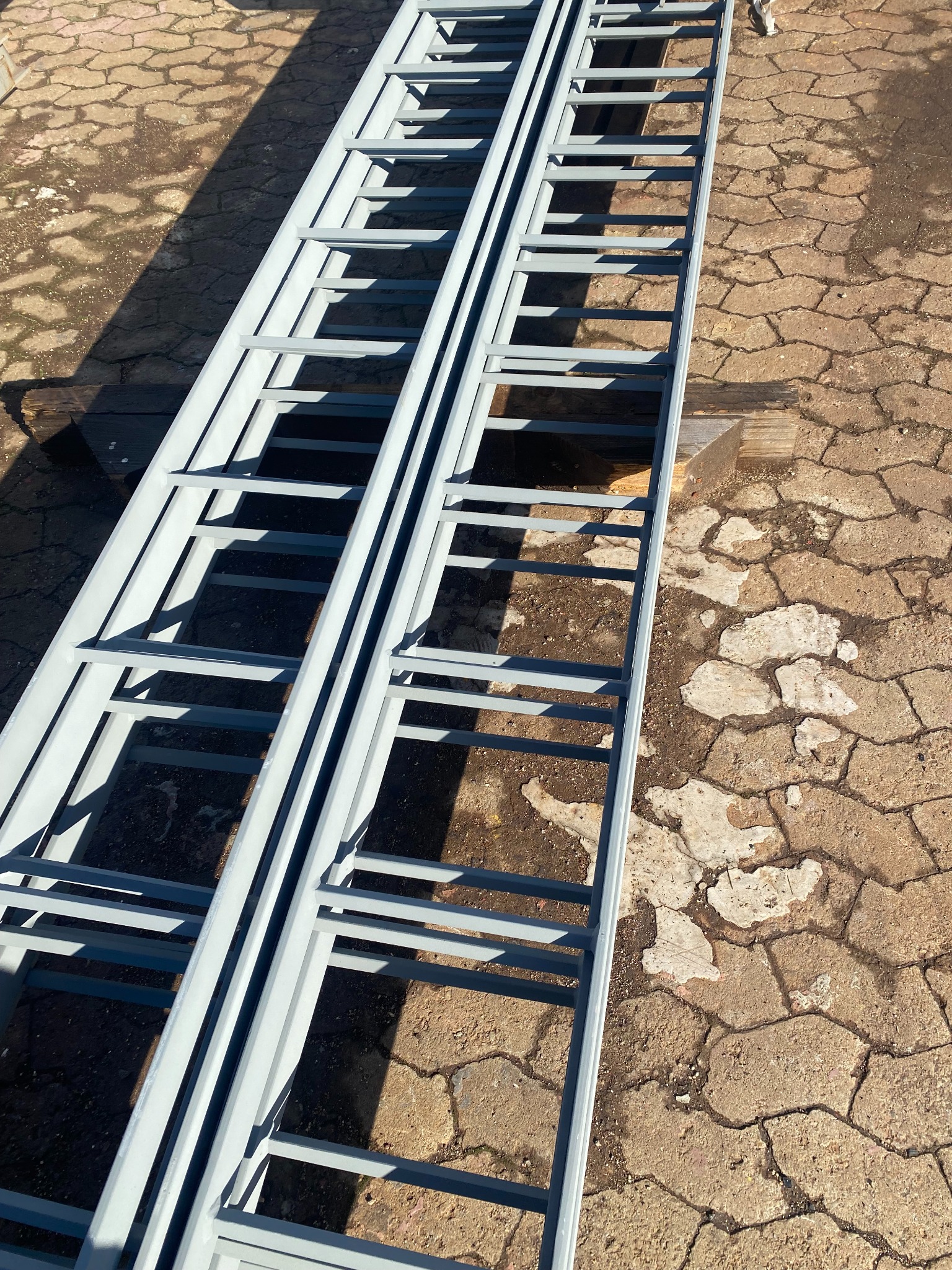 Dwie metalowe konstrukcje schodowe w kolorze szarym, przygotowane do montażu w Kartuzach, leżą na ziemi z betonowej kostki.