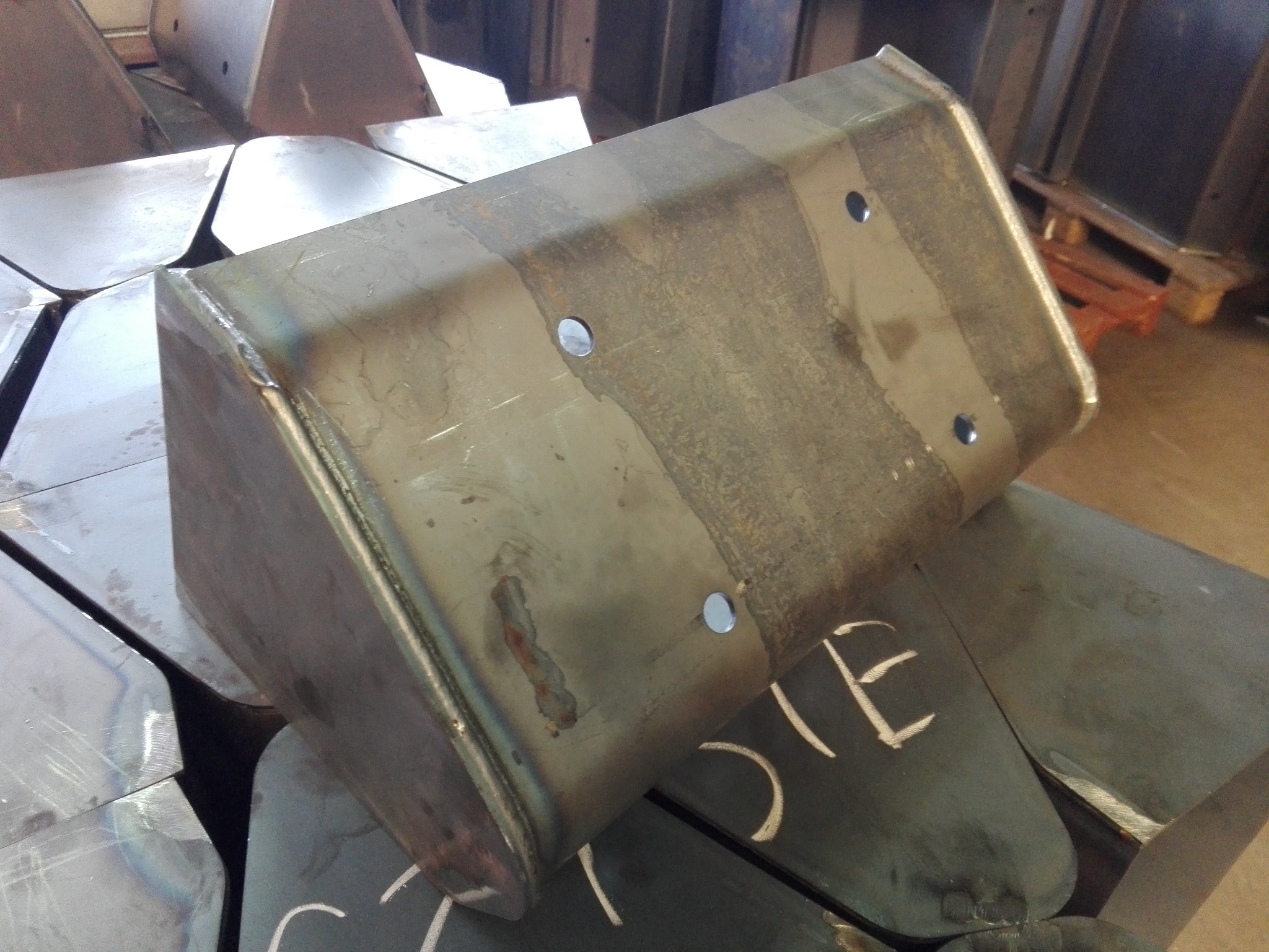 Metalowy element konstrukcyjny z widocznymi śladami spawania i otworami, ułożony na innych metalowych elementach z białymi oznaczeniami.