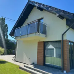 Dom z balkonem zabezpieczonym szkłem i nierdzewną stalą, widok z zewnątrz, jasna elewacja, ciemna dachówka, kostka brukowa i trawnik na pierwszym planie.