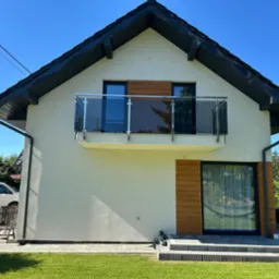 Nowoczesny dom z balkonem zabezpieczonym szklaną balustradą, widok z poziomu trawnika. Elementy wykończenia w odcieniach drewna, czarny dach i ramy okienne.