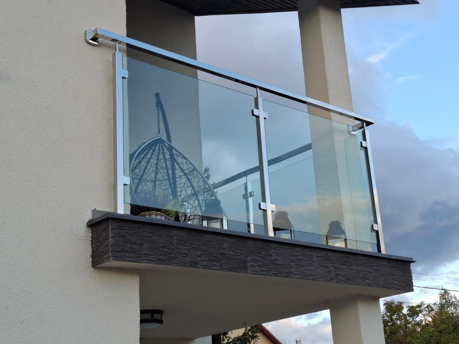 Szklana balustrada balkonowa z nierdzewnymi słupkami, widoczne odbicie wiszącego fotela kokon w szkle, elewacja budynku z dekoracyjną okładziną.