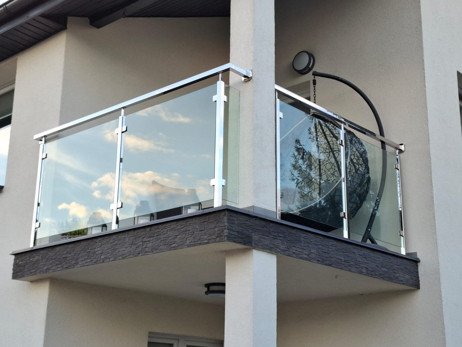 Balkon z nowoczesną balustradą ze szkła i stali nierdzewnej, w której odbijają się chmury, obok wiszące krzesło.