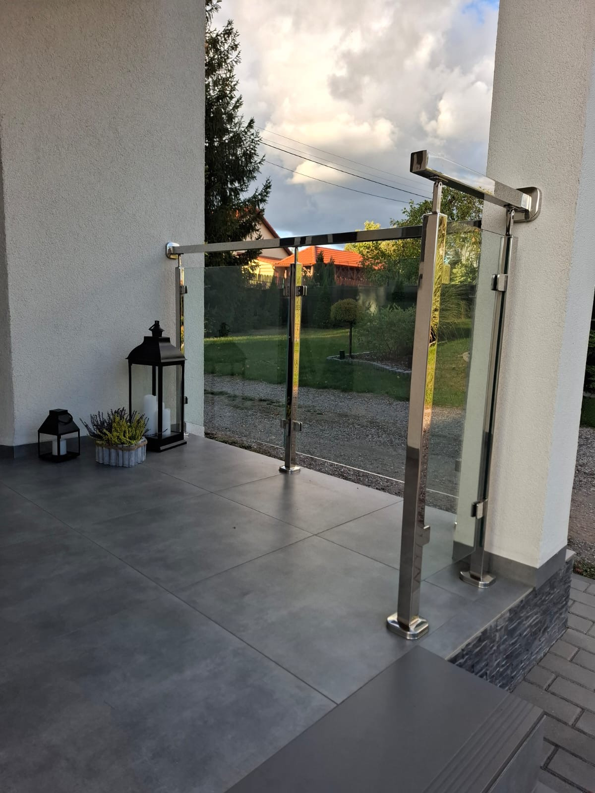 Szklana balustrada z polerowanymi słupkami ze stali nierdzewnej na tarasie, widok z zewnątrz, w tle dom z ogrodem.