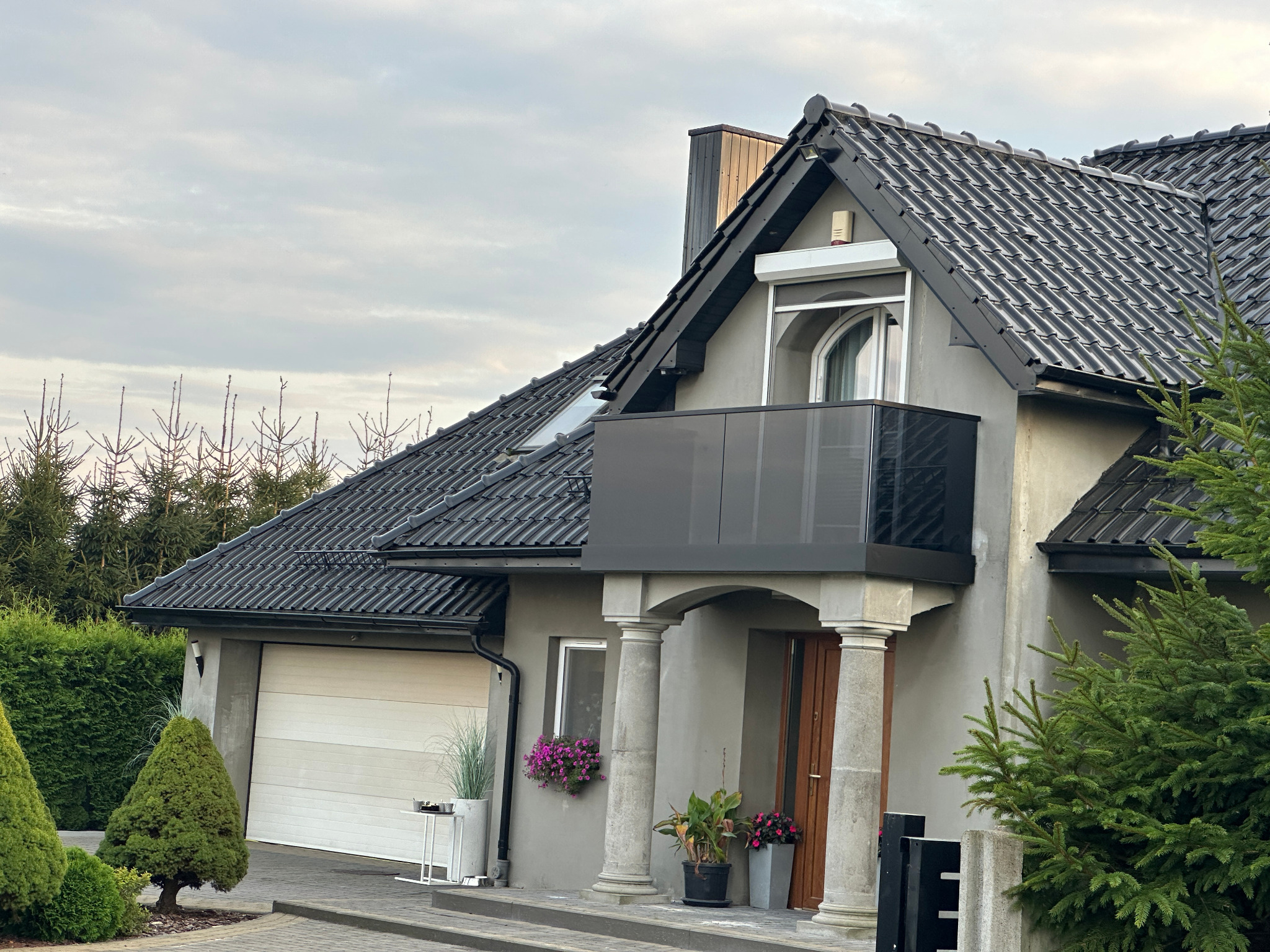 Nowoczesny dom jednorodzinny z balkonem zabezpieczonym szkłem, widoczne kolumny przy wejściu, szara elewacja i czarna dachówka, otoczony zielenią.