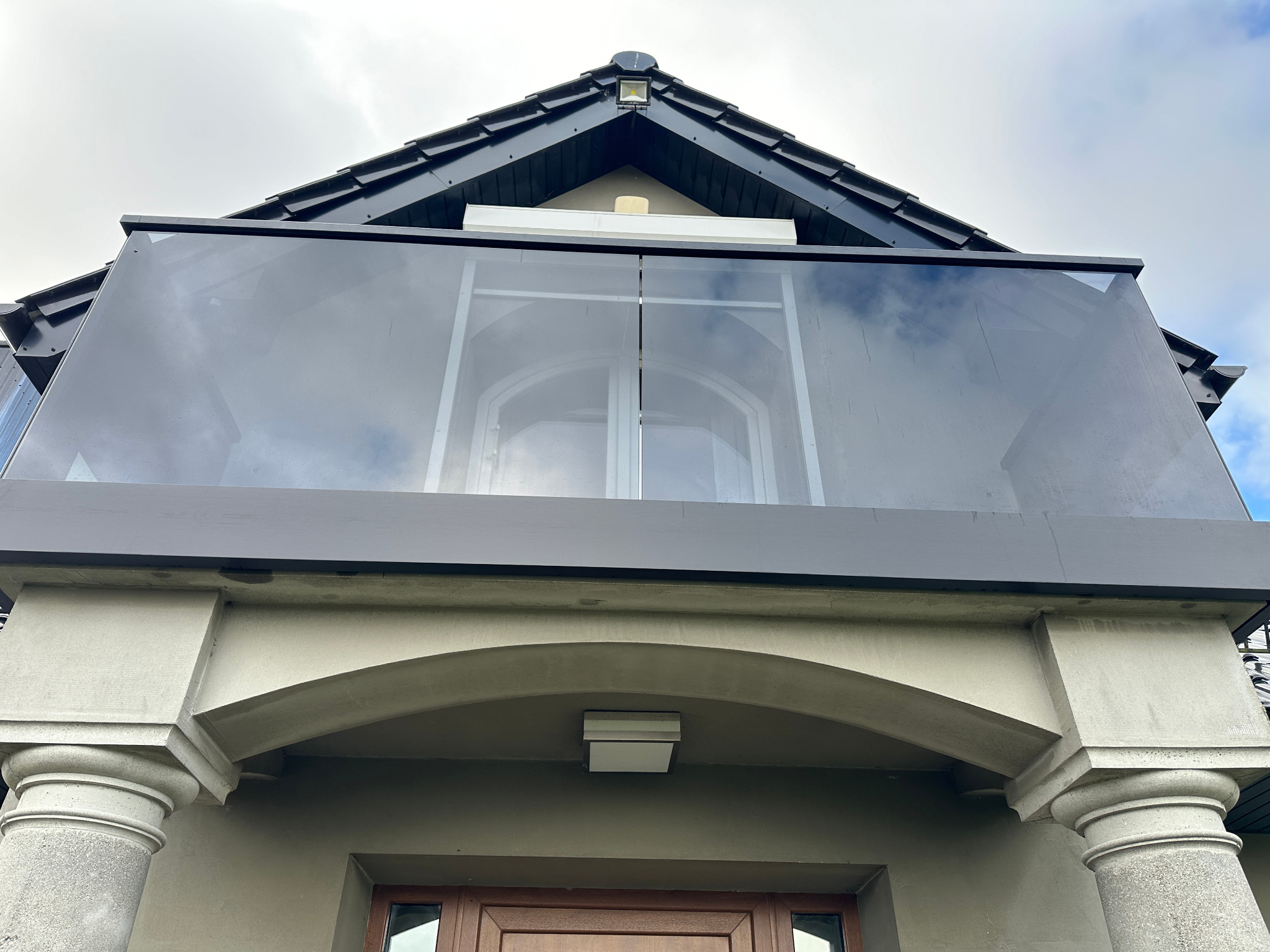 Szklana balustrada na balkonie z widokiem na pochmurne niebo, zamontowana na budynku z kolumnami i łukowym wejściem.