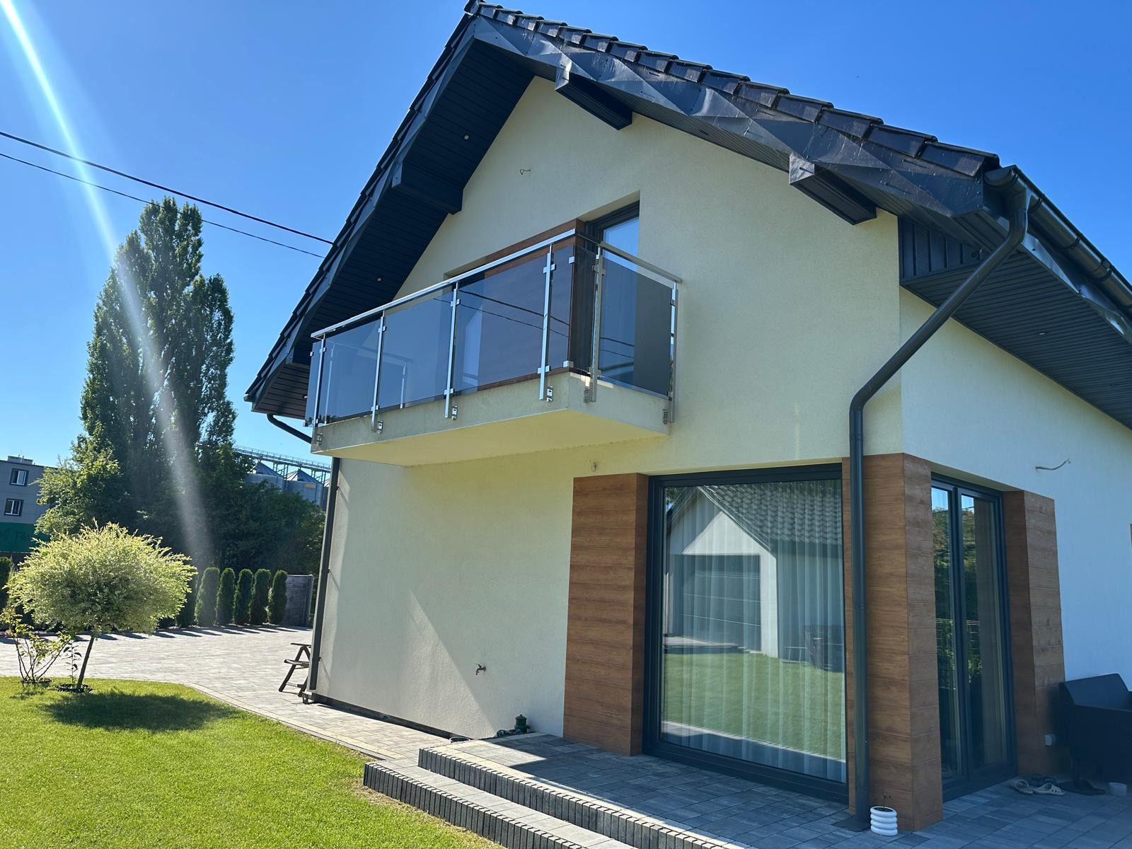 Dom z balkonem zabezpieczonym szkłem i nierdzewną stalą, widok z zewnątrz, jasna elewacja, ciemna dachówka, kostka brukowa i trawnik na pierwszym planie.